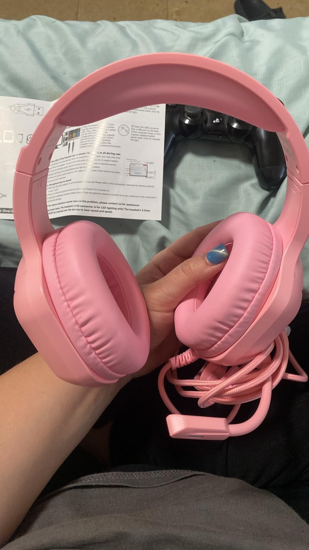 Pink IMYB A36 headset held in one hand with its braided cable coiled, instruction sheet and a PlayStation controller on a blanket behind