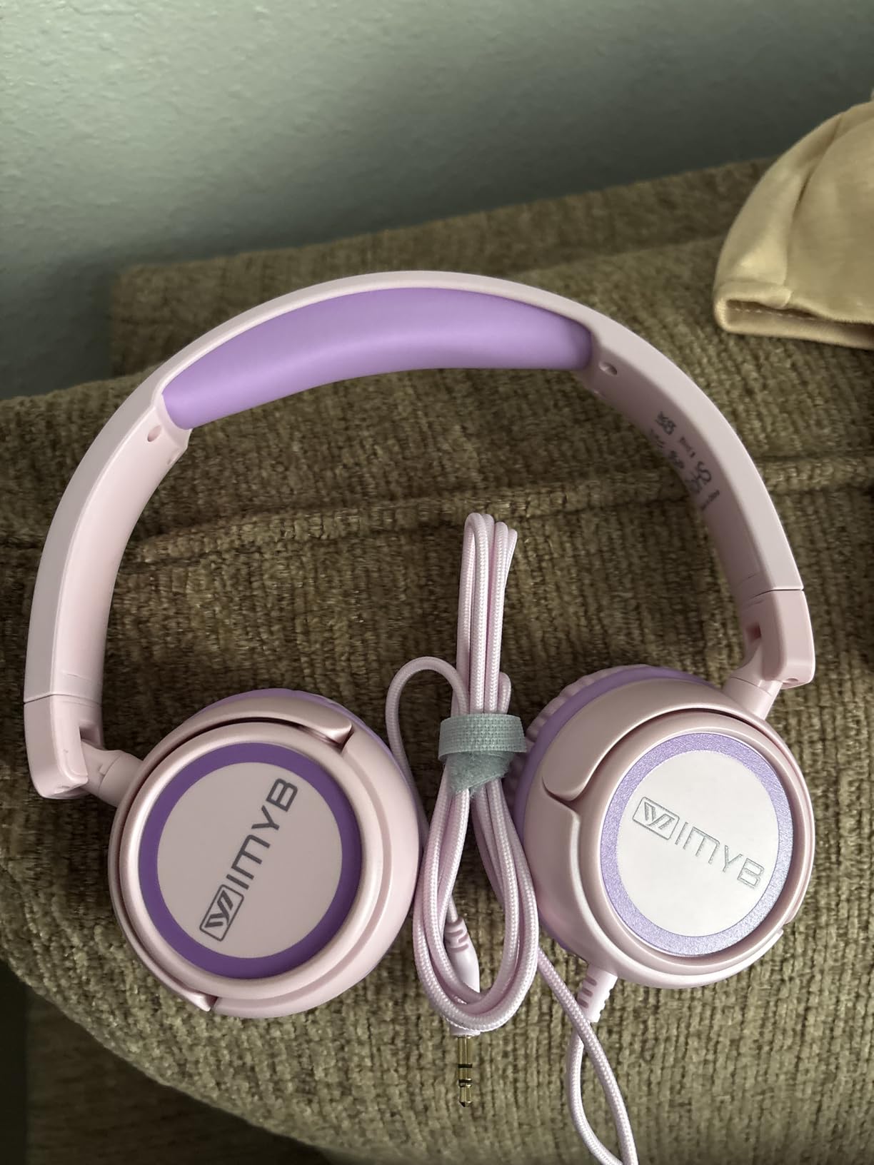 Pink and purple IMYB V1 kids headphones lying flat on a couch with the braided cable coiled and the 3.5mm plug visible