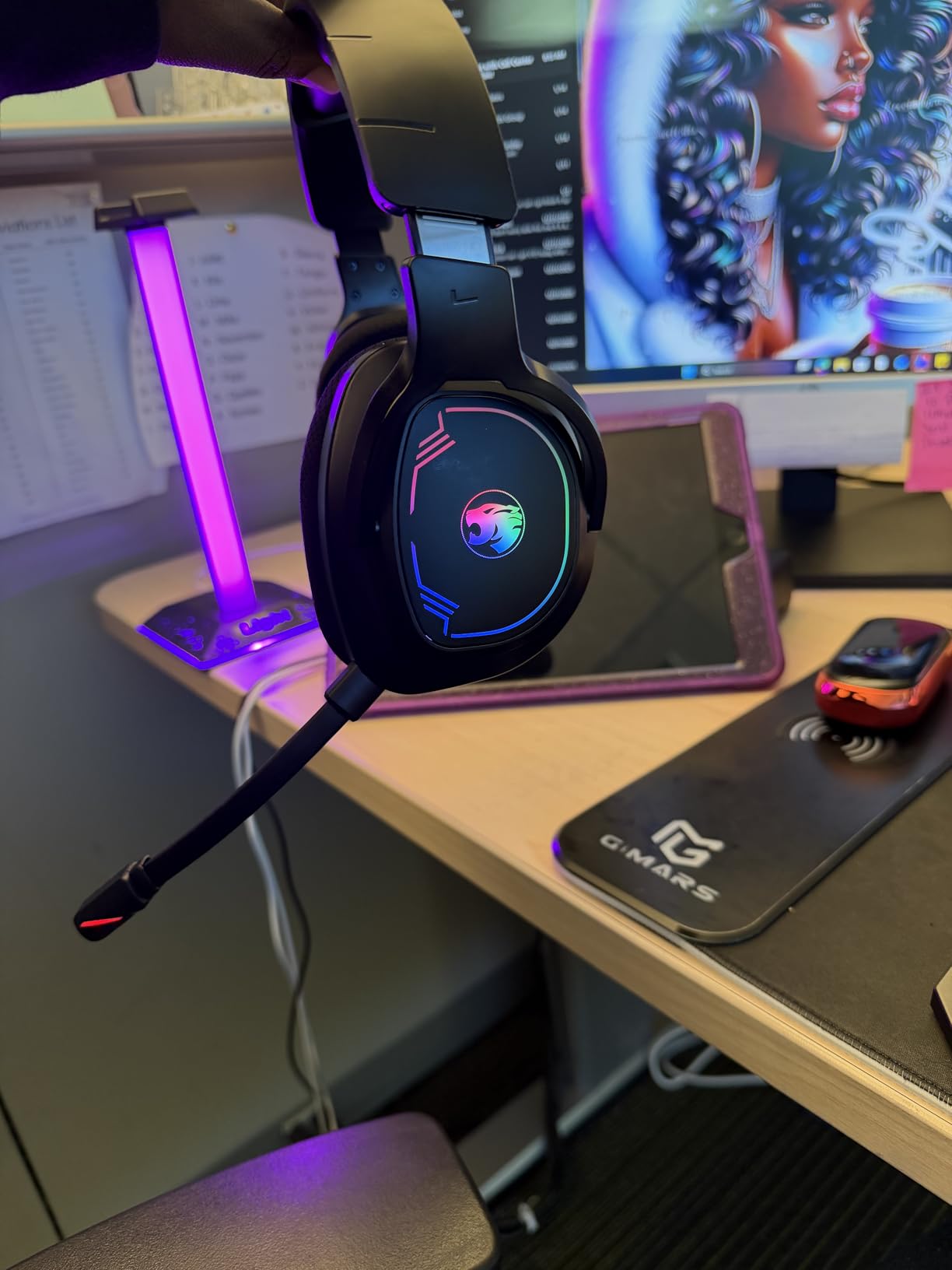 Black IMYB A36 headset held up beside a desk, RGB ring on the earcup lit green through blue to pink, microphone boom extended