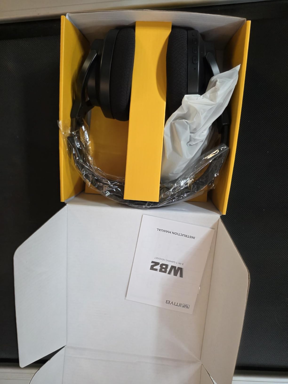 IMYB W82 headset unboxed in a yellow tray inside a shipping carton, with the 3-in-1 gaming headset instruction manual on the flap